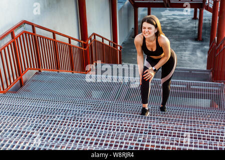 Portrait der junge sportliche Mädchen in schwarz Sportbekleidung stehen auf Treppen und haben starke verletzt Problem mit Knie, Spasmus schmerzhaft. Greifen Bein mit Hand mit Stockfoto