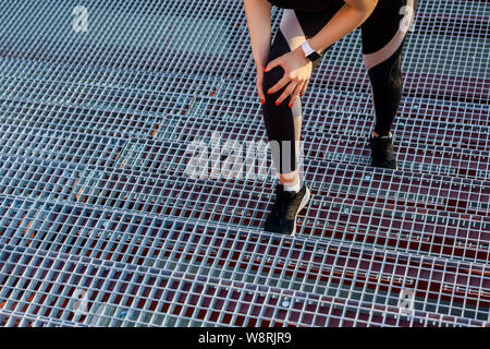 Closeup Portrait von junge sportliche Mädchen in schwarz Sportbekleidung stehen auf Treppen und haben eine starke verletzt Problem mit Knie, Spasmus schmerzhaft. Greifen Bein mit Stockfoto