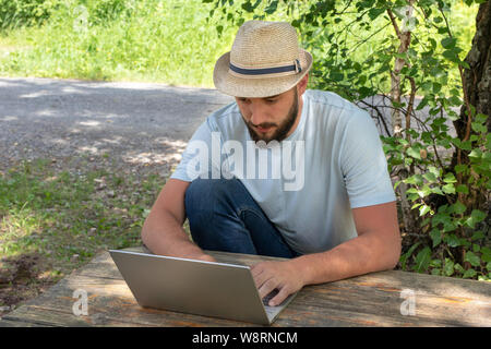Man ist bemüht, einen Computer an einem Tisch im Park. Das Konzept der remote arbeiten, Büro, freiberufliche Stockfoto