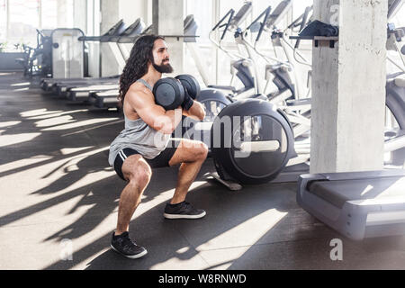 Portrait von starken jungen erwachsenen Mann mit langen Locken schöner Athlet im Fitnesscenter zu trainieren, statische Hocke und halten Sie zwei Kurzhanteln, Übungen f Stockfoto