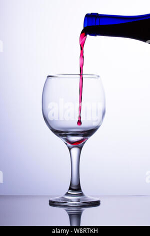 Der erste Tropfen fällt in ein leeres Glas. Ein Jet der roten Wein fließt in ein Glas aus einer blauen Flasche. Wein Jet nicht das Glas erreichen. Stockfoto