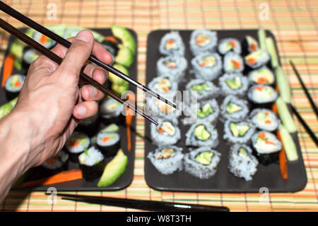 Hand mit Stäbchen mit Hintergrund voll von verschiedenen Arten von Sushi zu essen. Die Auswahl und das Essen von traditionellen Sushi mit japanischen Dekoration. Stockfoto