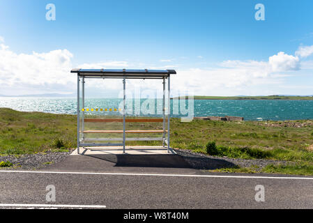 Leeren Bus Stop mit einem Glas Unterschlupf in einer abgelegenen Lage entlang einer Straße die Küste an einem klaren Sommertag. Malerische Bucht im Hintergrund. Stockfoto