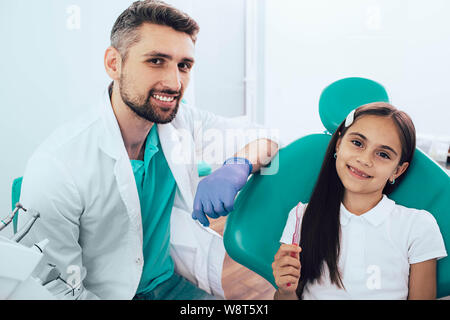 Wenig gemischten Rennen Mädchen im zahnmedizinischen Stuhl in der Nähe von Ihrem Zahnarzt sitzen. Sie holding Zahnbürste und lächelnd. Die richtige Bürste Zähne Stockfoto