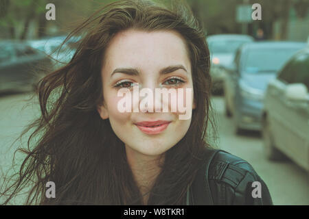 Happy girl außerhalb posiert in der Straße mit parkenden Autos auf den Hintergrund auf Kamera mit windigen Haar Stockfoto