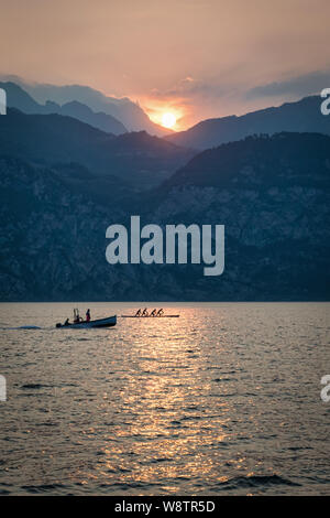 Dies ist ein Sonnenuntergang Bild des Gardasees. Im Vordergrund steht ein Fischerboot und ein Ruderboot Team. Im Hintergrund ist die Sonne hinter den Stockfoto