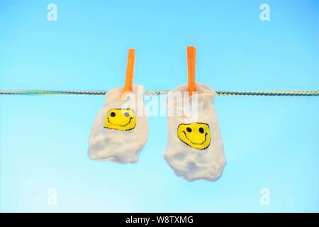 Zwei weiße Socken mit einem gelben Smiley Emoticon hängen an einem Seil mit Wäscheklammern, blauer Himmel Hintergrund Stockfoto