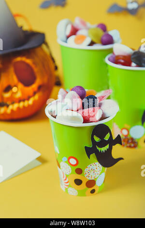 Halloween Trick oder Süßigkeiten zu behandeln. Pappbecher mit bunten Bonbons innen und Papier Silhouetten der Fledermäuse, Geister und Hexe auf gelben Hintergrund. Stockfoto