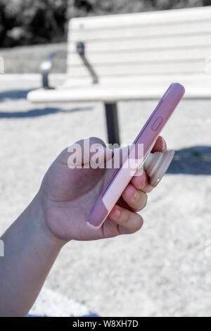 Hand hält eine Frau ein Smartphone in einem rosa gegen eine de-gesättigten Hintergrund mit einer Parkbank. Stockfoto