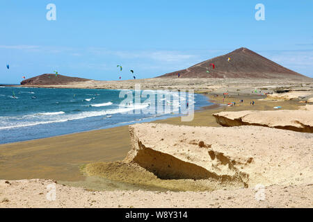 Viele bunte Drachen am Strand und Kitesurfer, Wellen und Fliegen bei windigen Tag im kanarischen El Medano auf Teneriffa mit Montana Roja Hügel auf Ho Stockfoto