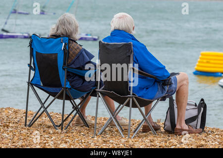 Ein älteres grauhaariges Paar, das an einem windigen Tag in zusammenklappbaren Sitzplätzen am Strand am Meer sitzt. Stockfoto