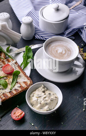 Leckeres Frühstück. Toast mit Frischkäse und Rucola und Cappuccino auf einem dunklen Hintergrund. Vertikaler Dampfstoß Stockfoto