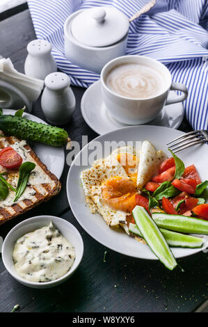 Leckeres und gesundes Frühstück. Gebratene Eier, Salat, Toast mit Frischkäse und Rucola und Cappuccino auf einem dunklen Hintergrund. Vertikaler Dampfstoß Stockfoto