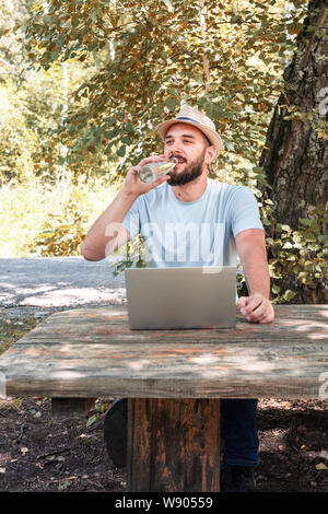 Junger Mann mit Bart, hipster Durst stillt, Getränke Wasser aus einer Flasche beim Sitzen an einem Tisch im Park Stockfoto