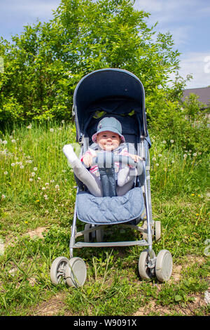 Ein 9 Monate altes Mädchen sitzt in einem Sommer Schlitten und spielt mit ihren Beinen. Wenig lächelnde Kind in einem Hut auf einem Spaziergang, sonnigen Sommertag Stockfoto