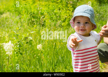 Kleines Mädchen, 9 Monate alt, Spaziergänge in einer Wiese mit ihren Eltern. Portrait von ein lächelndes Kind in Panama und Kleid. Das Mädchen hält die Hand. Baby Stockfoto