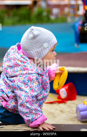 Baby Mädchen ein Jahr alt in warmen demi-Saison Kleidung Hut Jacke Wanderungen spielt in der Sandbox. Ein Kind spielt auf dem Spielplatz. Mädchen spielen im Sand Dosen, Stockfoto