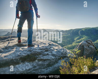 Man touristische mit festen Bein macht Schritt auf felsigen Hügel mit Hilfe des Unterarms Krücken. Stockfoto