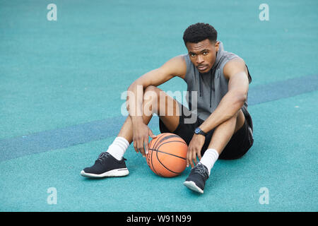 Volle Länge Portrait von Stattlichen afrikanische Sportler sitzen auf dem Boden im Basketball Court und Kamera, kopieren Raum Stockfoto