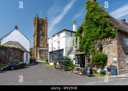 Cerne Abbas, England, UK - 29. Juni 2019: ein Mann Getränke außerhalb der Royal Oak Pub in der Ortschaft Cerne Abbas, mit der Kirche St. Mary hinter sich. Stockfoto