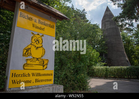 Zossen, Deutschland. 06 Aug, 2019. Ein Schild mit der Aufschrift 'Bücher und Bunker Stadt Wünsdorf "begrüßt die Besucher des Dorfes. Rechts im Bild steht eine hohe Bunker vom Typ Winkel. Der Bunker Stadt Wünsdorf, etwa 40 Kilometer von Berlin entfernt, ist einer der wichtigsten Standorte in der Geschichte des nationalsozialistischen Deutschland. Credit: Monika Skolimowska/dpa-Zentralbild/ZB/dpa/Alamy leben Nachrichten Stockfoto