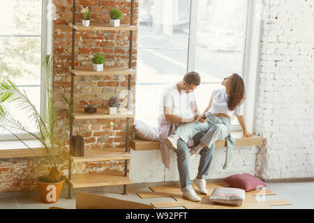Junges Paar zog in ein neues Haus oder Wohnung. In der Nähe von Fenster mithilfe von Smartphone und entspannend nach der Reinigung und auspacken. Glücklich und zuversichtlich. Familie, Verschieben, Beziehungen Konzept. Stockfoto