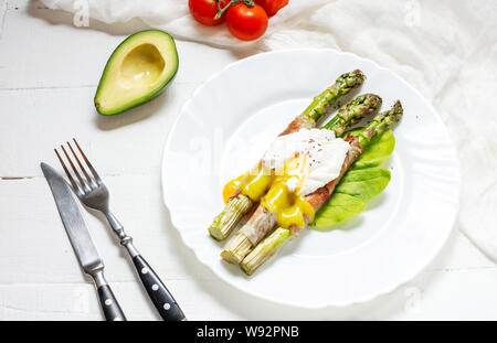 Gegrillter grüner Spargel mit Speck umwickelt, Benedikt pochiertes Ei auf weißem Holz Hintergrund. Frühstück Stockfoto