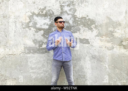 Dies ist mir. Portrait von Stolz gut aussehender bärtiger junger Mann in karierten blaues Hemd und Sonnenbrille gegen die konkrete graue Wand stehend. angezeigt selbst ein Stockfoto