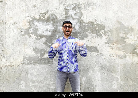 Wer? Mich? Portrait von überrascht gut aussehender bärtiger junger Mann in karierten blaues Hemd und Sonnenbrille gegen die konkrete graue Wand stehend, sich zeigen Stockfoto