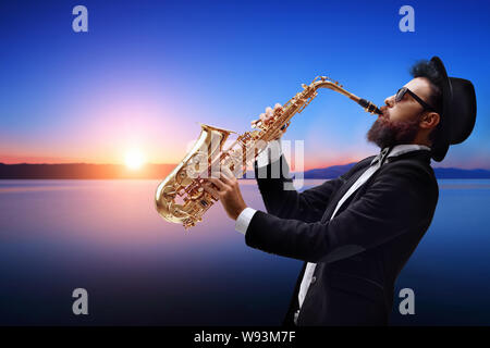 Männliche Jazz Musiker spielen ein Saxophon mit einem Sonnenuntergang und Wasser hinter ihm Stockfoto