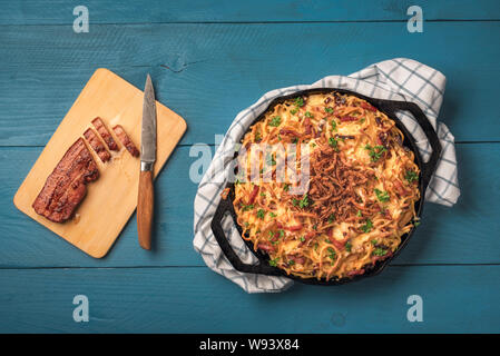 Bayerische Nudeln mit Käse, Speck und gebratene Zwiebel in der Pfanne Gusseisen, auf blauen Holztisch. Oben Blick auf traditionelle deutsche Küche. Stockfoto