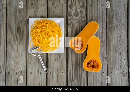 Overhead Blick von oben auf die frisch geschälten Kürbis Nudeln und Julienne Schäler. Holz- Hintergrund und weiße Platte für frische, natürliche pflanzliche Nudel Stockfoto