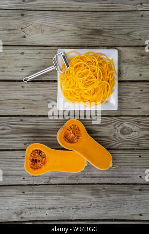 Overhead Blick von oben auf die frisch geschälten Kürbis Nudeln und Julienne Schäler. Holz- Hintergrund und weiße Platte für frische, natürliche pflanzliche Nudel Stockfoto