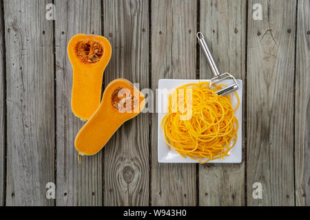Overhead Blick von oben auf die frisch geschälten Kürbis Nudeln und Julienne Schäler. Holz- Hintergrund und weiße Platte für frische, natürliche pflanzliche Nudel Stockfoto