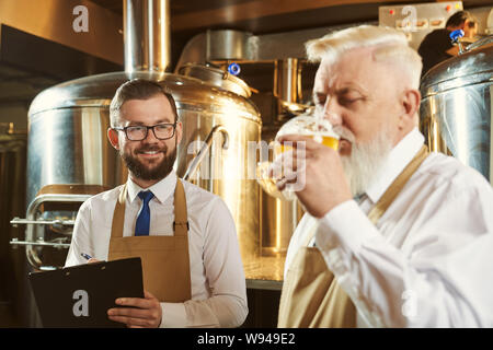 Selektiver Fokus der glückliche junge Brauer, Ordner, lächeln und Kollegen. Mann, Glas Bier und köstliche golden Ale. Begriff der Produktion und trinken. Stockfoto