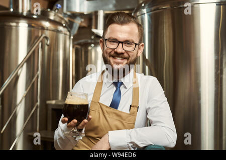 Vorderansicht der freundliche Mann mit weißem Hemd und Schürze auf Kamera und lachend, während Sie ein Glas Bier. Professionelle Brauer genießen Leckere dunkle Ale. Begriff der Produktion. Stockfoto
