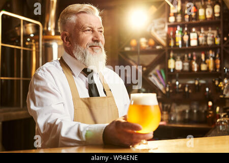 Blick auf glückliche männliche Brauer stehend über Bar 7/8 und Glas goldenen Bier in der Hand. Mann im weißen Hemd und Schürze freuen und Lächeln. Konzept der ALE-Produktion. Stockfoto