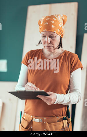 Frau Carpenter tragen Bandanna schreibt Projekt Hinweise zur Zwischenablage beachten Sie Papier in ihrem kleinen Geschäft Holzarbeiten workshop Stockfoto