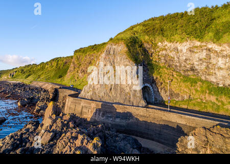 Black Arc Tunnel und Causeway Coastal Route. Malerische Straße entlang der östlichen Küste des County Antrim, Nordirland, Großbritannien. Luftaufnahme im Sonnenaufgang Licht Stockfoto