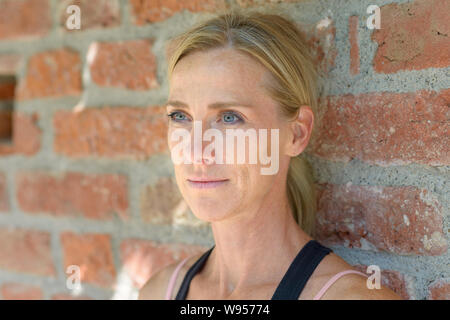 Portrait von eine wunderschöne blonde junge Frau auf der Suche nach mit ihren blauen Augen beim Tagträumen über Zukunft gegen eine Mauer im Freien im Sommer Stockfoto