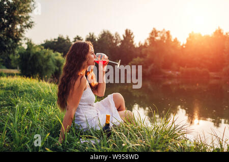 Junge Frau mit einem Glas Wein am Ufer bei Sonnenuntergang. Frau Landschaft bewundern, während Sie trinken im Sommer Park Stockfoto