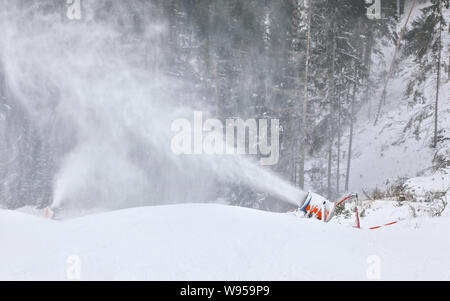 Orange und Blau Beschneiung Cannon ausbreitenden Eiskristalle über Piste, Bäume im Hintergrund Stockfoto