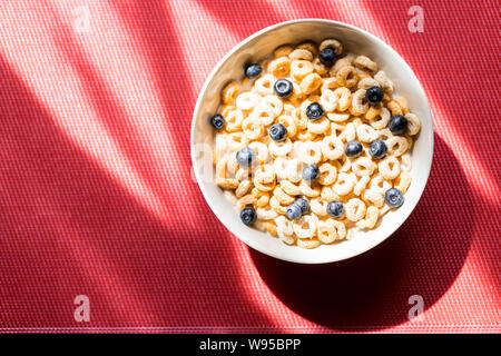 Gesundes Frühstück mit Milch, Müsli und Obst auf dem roten Tisch. Weizen Flocken mit Blaubeeren gemischt, in einem weißen Keramik Schüssel serviert für einen gesunden Stockfoto