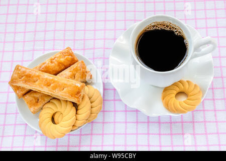 Süße Blätterteig cookies auf einem weißen Teller und Tasse mit heißem Kaffee schwarz auf weiß rosa kariert Tischdecke. Leckeres Frühstück oder einen Snack. Stockfoto