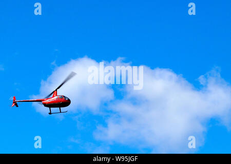 Ein kleiner roter Hubschrauber fliegt durch den blauen Himmel mit weißen Wolken. Luftverkehr. Stockfoto