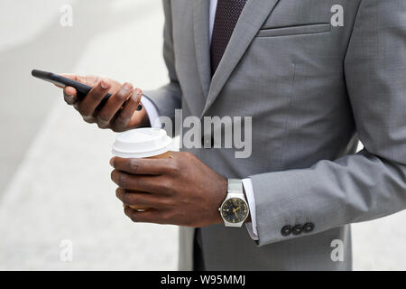 Nahaufnahme der Afrikanischen Geschäftsmann in Anzug holding Tasse Kaffee und über Handy für seine online Arbeiten im Freien Stockfoto