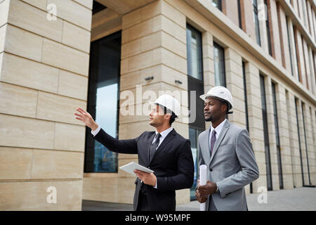 Der junge Architekt in hardhat zeigen auf Gebäude und erklären das neue Projekt zu seinen afrikanischen Kollegen draußen Stockfoto