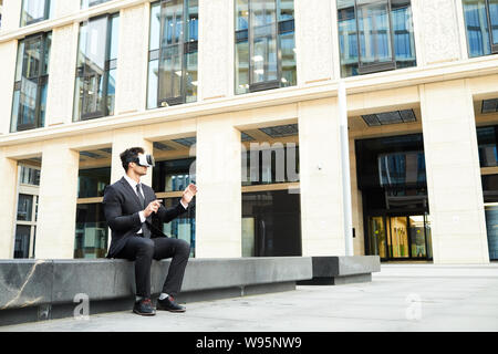 Junge Unternehmer mit Virtual reality Brillen online Kommunikation beim Sitzen im Freien in der Nähe des Bürogebäudes Stockfoto