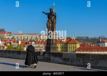 Bischof oder Priester zu Fuß in der Nähe der Statue des Heiligen Johannes des Täufers auf Sockel und Balustraden der Karlsbrücke über die Moldau in sonniger Tag. Stockfoto