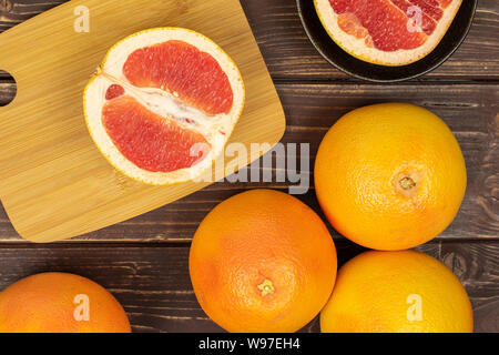 Gruppe von vier ganze zwei Hälften von frischem rosa Grapefruit in dunklen Keramik Schüssel auf Bambus Schneidebrett flatlay auf braunem Holz Stockfoto